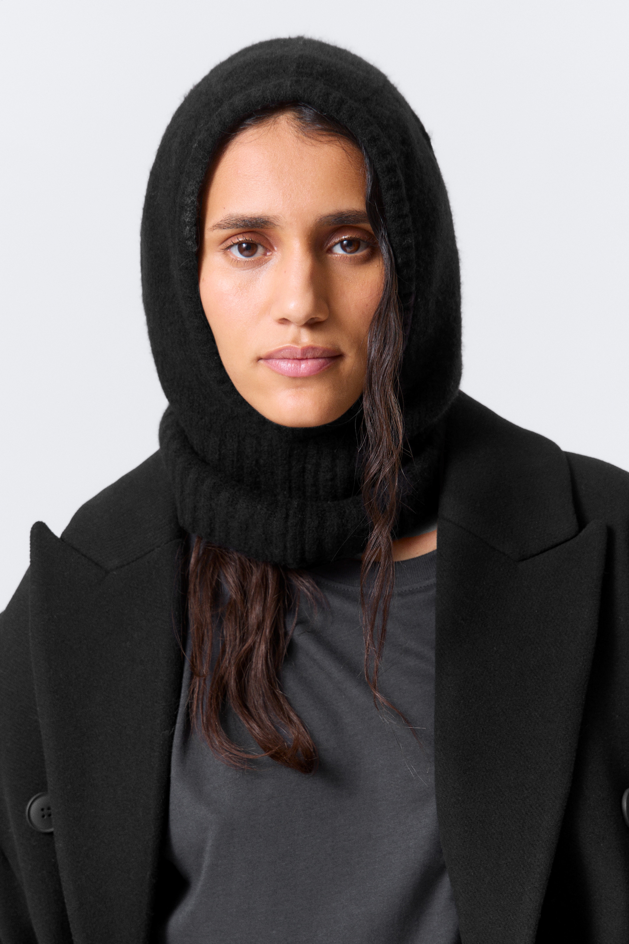 Grösseres Bild ansehen: Eine schwarze gestrickte Kapuze wird getragen, aus einer Frontalansicht präsentiert. Sie bedeckt den Kopf und Hals, umrahmt das Gesicht eng mit einer weichen Strickstruktur am Kopf und einem gerippten Strick, der sich um den unteren Hals erstreckt. Die Kapuze ist über einem grauen Hemd und unter einem schwarzen Mantel geschichtet.