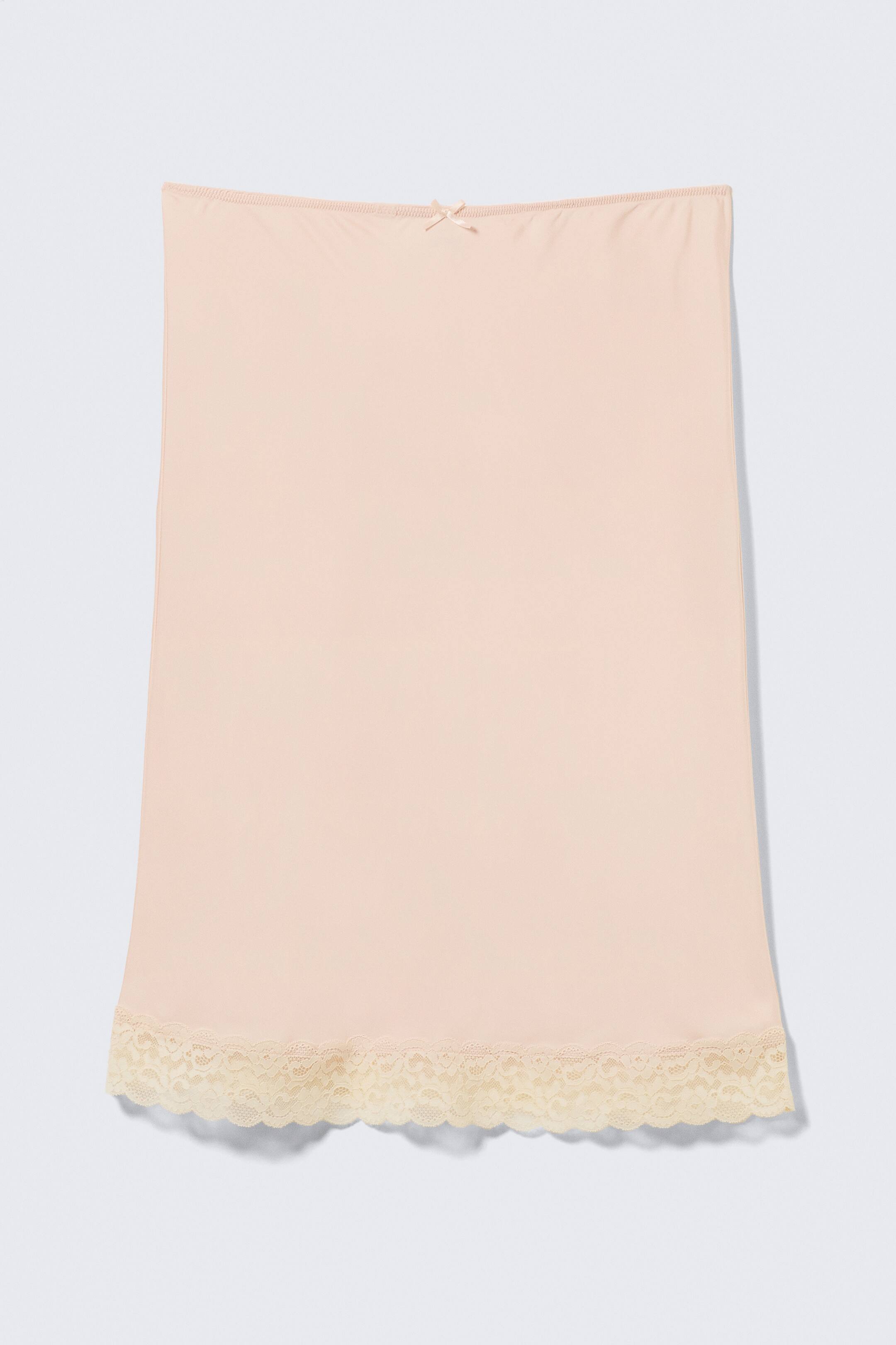 Visualizza immagine più grande: Una gonna midi con bordo in pizzo è presentata distesa, fronte frontale. È in raso color pesca chiaro, caratterizzata da una fascia elastica in vita con un piccolo fiocco centrale in raso e un bordo in pizzo intricato sull'orlo inferiore in una tonalità leggermente più chiara.