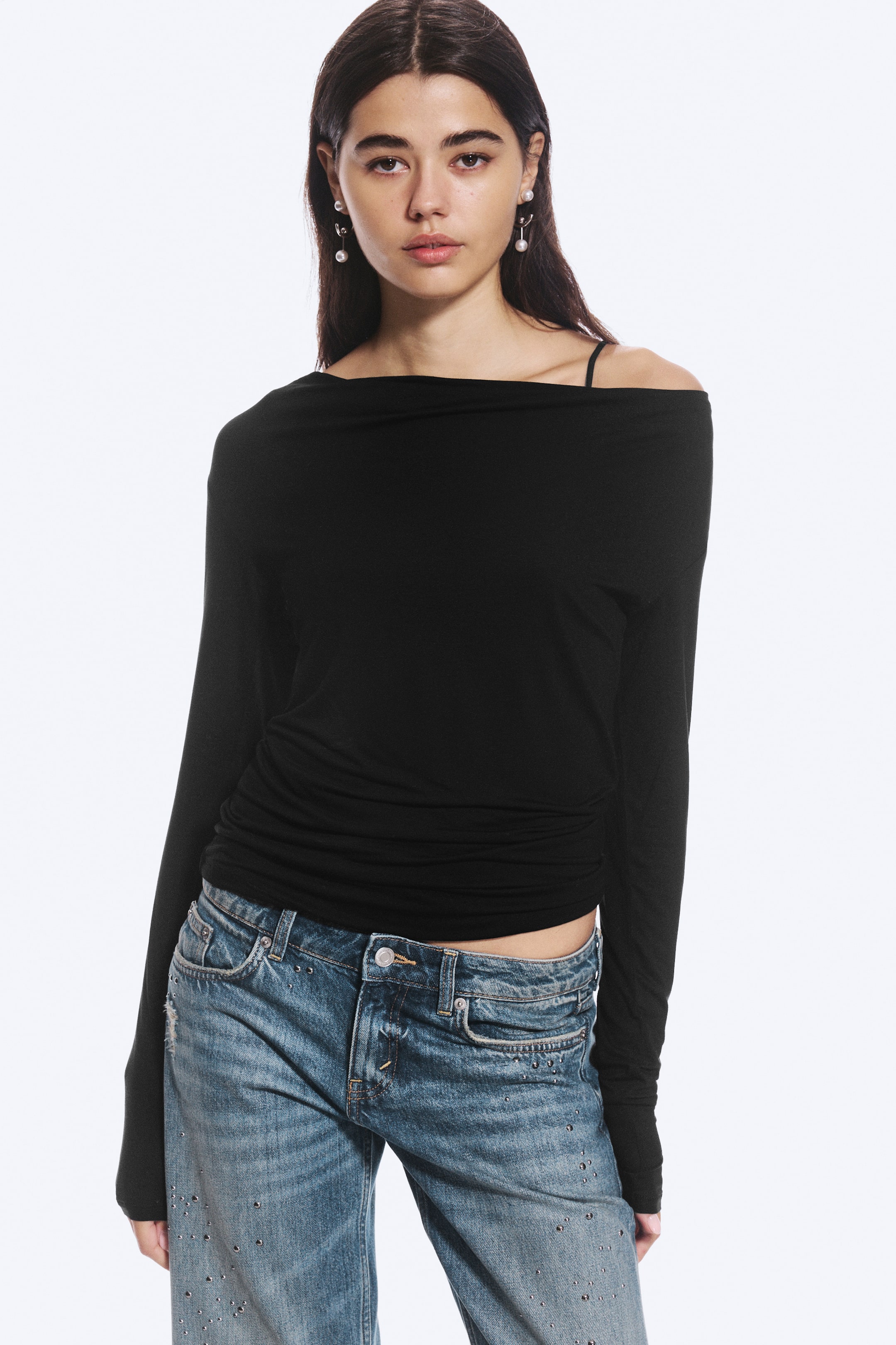 Visualizza immagine più grande: La modella indossa un top nero slim fit a maniche lunghe, caratterizzato da uno scollo a cowl drappeggiato e asimmetrico e da dettagli arricciati in vita. Il top è abbinato a jeans in denim blu.