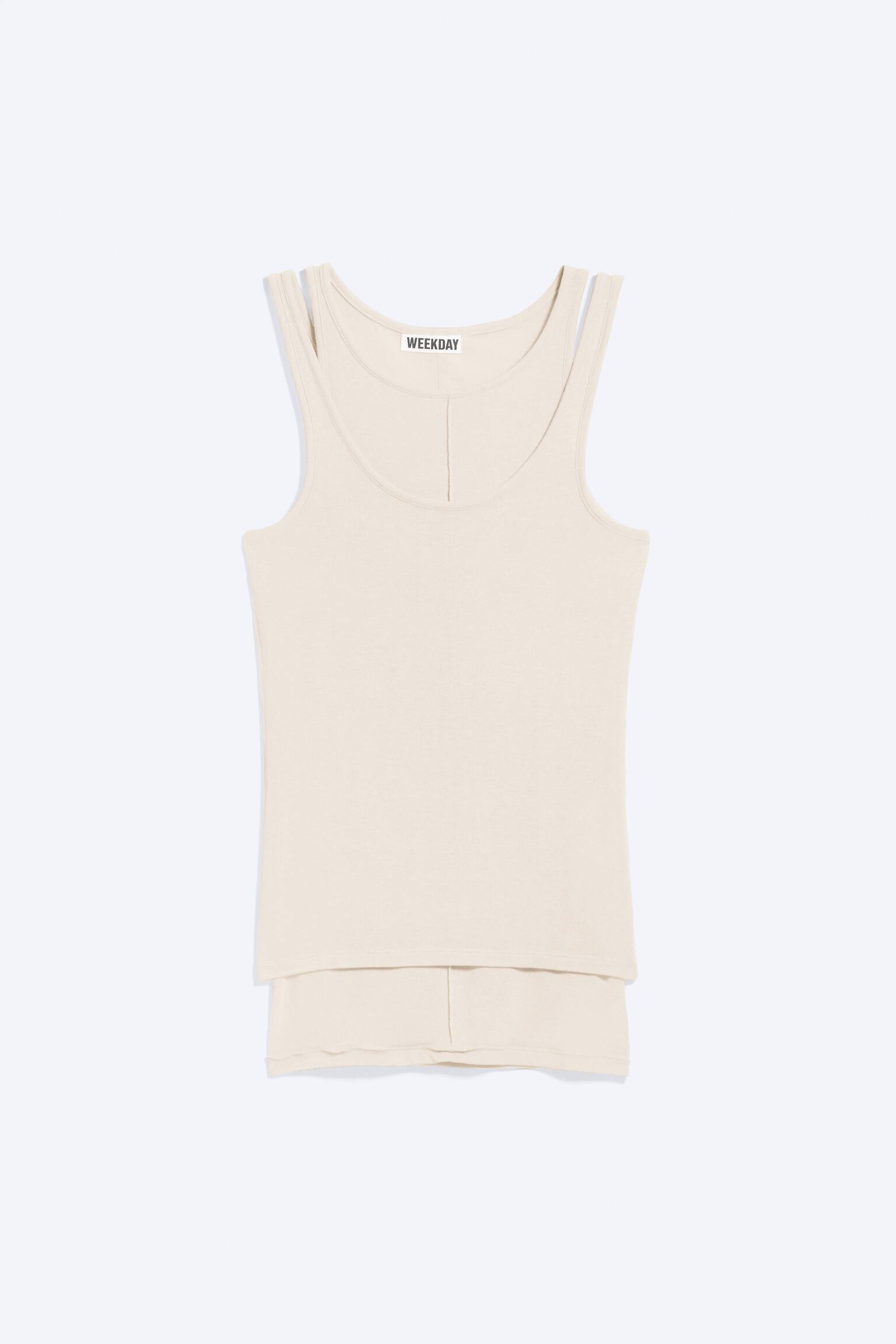 Una camiseta de tirantes color crema presenta su vista frontal, con un escote redondo y una etiqueta de la marca 'WEEKDAY' visible en el interior del cuello. Se aprecian dobles tirantes finos y una costura vertical sutil que recorre el centro del frente. La camiseta de tirantes está colocada sobre un artículo de tela a juego en la parte inferior. - 1