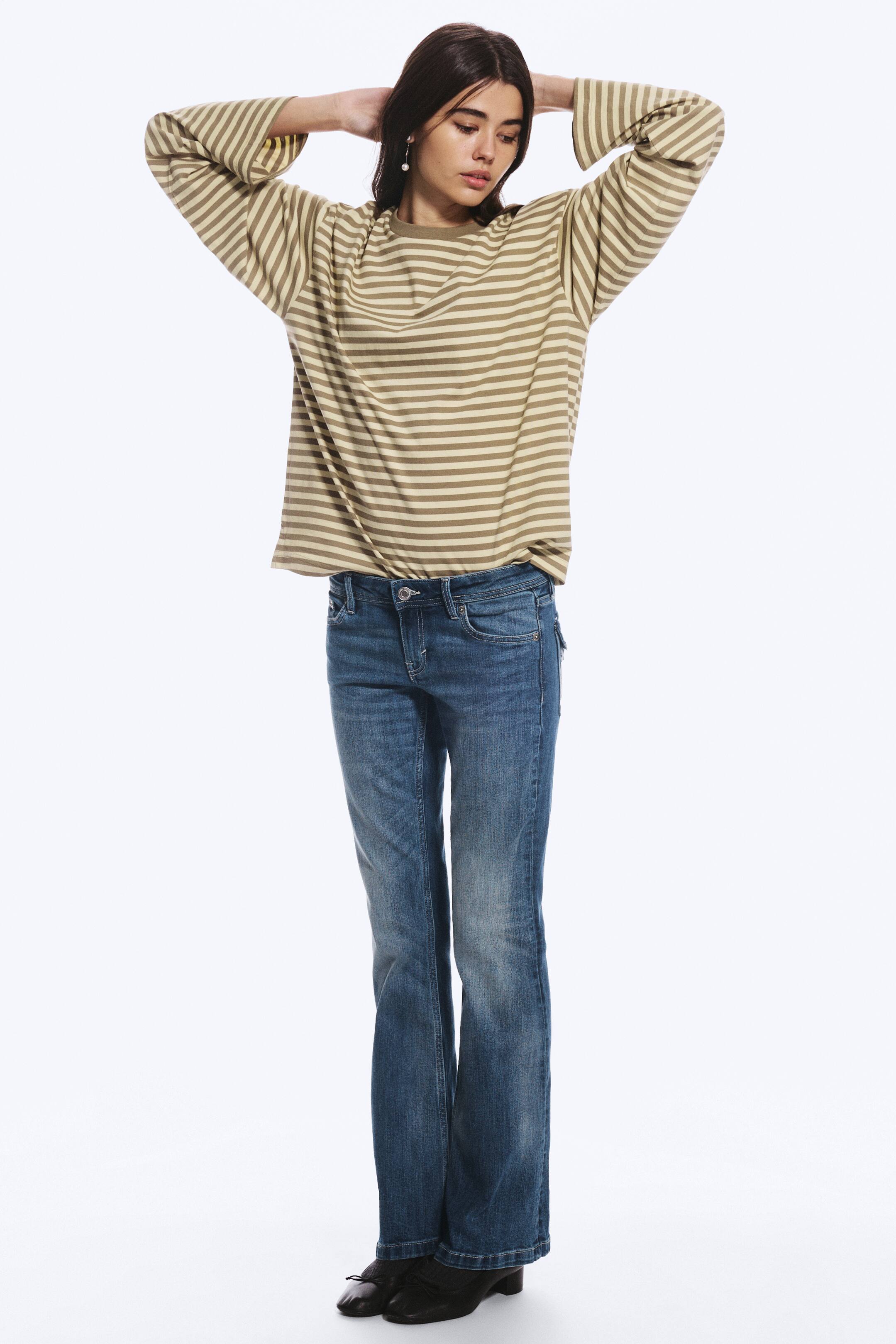 Agrandir l'image: Un jean bootcut taille basse, en délavage bleu moyen, porté par le mannequin et associé à un haut à manches longues rayé. Le jean présente des poches avant, des passants de ceinture et une coupe regular sur toute la jambe.