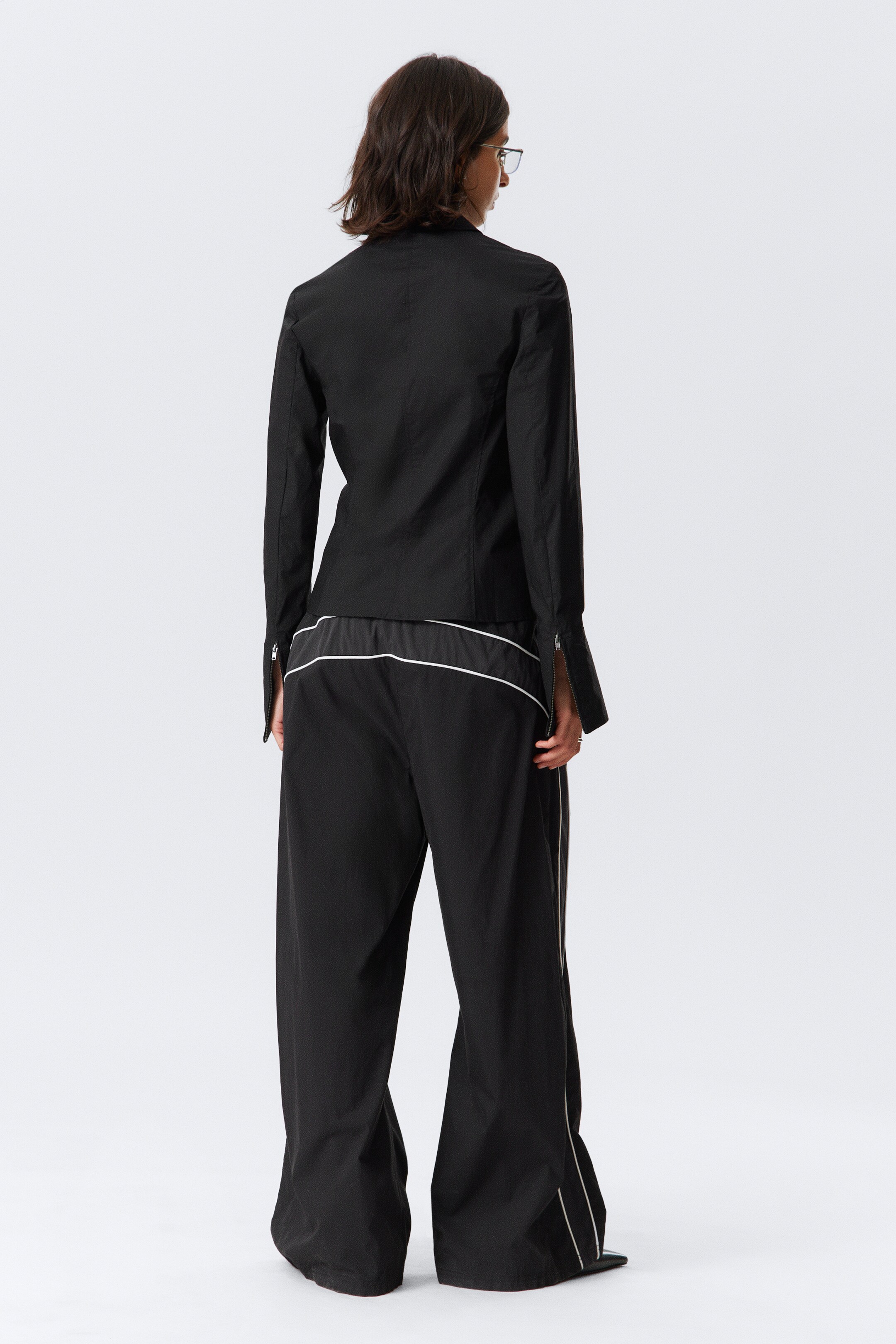 Visualizza immagine più grande: Una persona sta indossando un top nero strutturato con maniche lunghe e pantaloni neri a gamba larga, presentati da una vista posteriore. Il top presenta polsini con cerniera parzialmente aperti, mentre i pantaloni a vestibilità rilassata sono dettagliati con una bordatura bianca lungo le cuciture laterali e una striscia bianca sulla cintura.