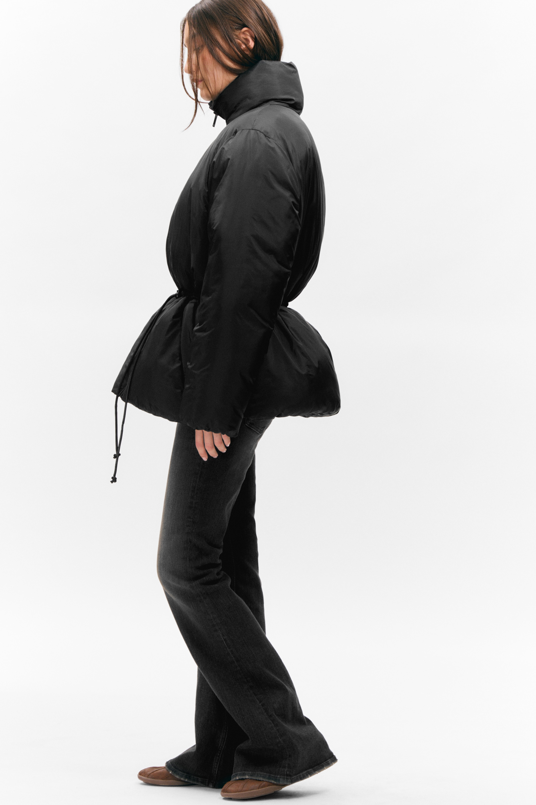 Agrandir l'image: Une doudoune noire est vue de profil, avec un col montant, des manches longues et bouffantes, et une taille resserrée pour former un ourlet en style péplum volumineux et froncé, avec un cordon de serrage visible.
