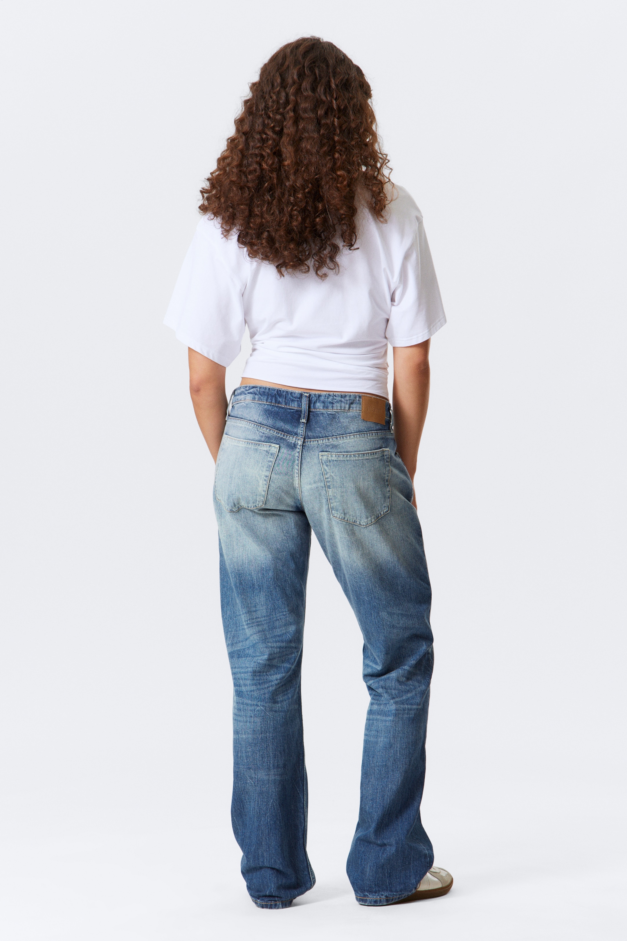 Visualizza immagine più grande: Un paio di jeans a lavaggio blu medio viene mostrato da dietro, caratterizzato da una vestibilità rilassata e gamba dritta. I jeans presentano dettagli strappati sulle cosce e sul sedile, insieme a due tasche posteriori a toppa visibili e una toppa in similpelle marrone sulla cintura.