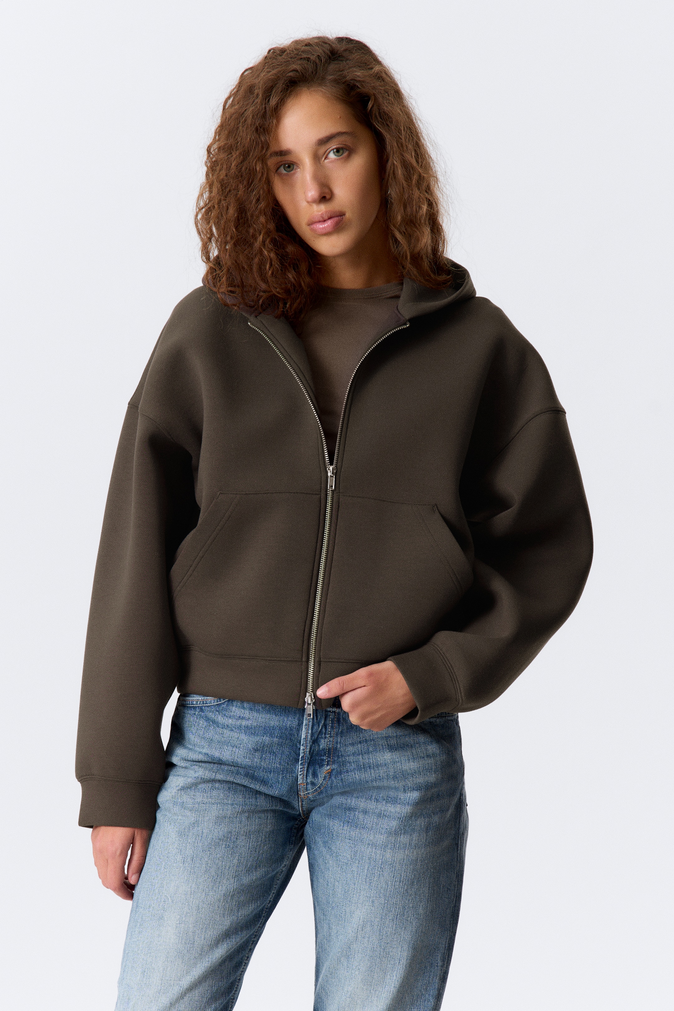 Agrandir l'image: Un sweat à capuche zippé de couleur vert olive foncé est porté face avant, avec une fermeture éclair argentée sur le devant, une capuche et deux poches avant. Il est conçu avec une longueur courte et est superposé à un haut à col rond assorti de couleur vert olive foncé.