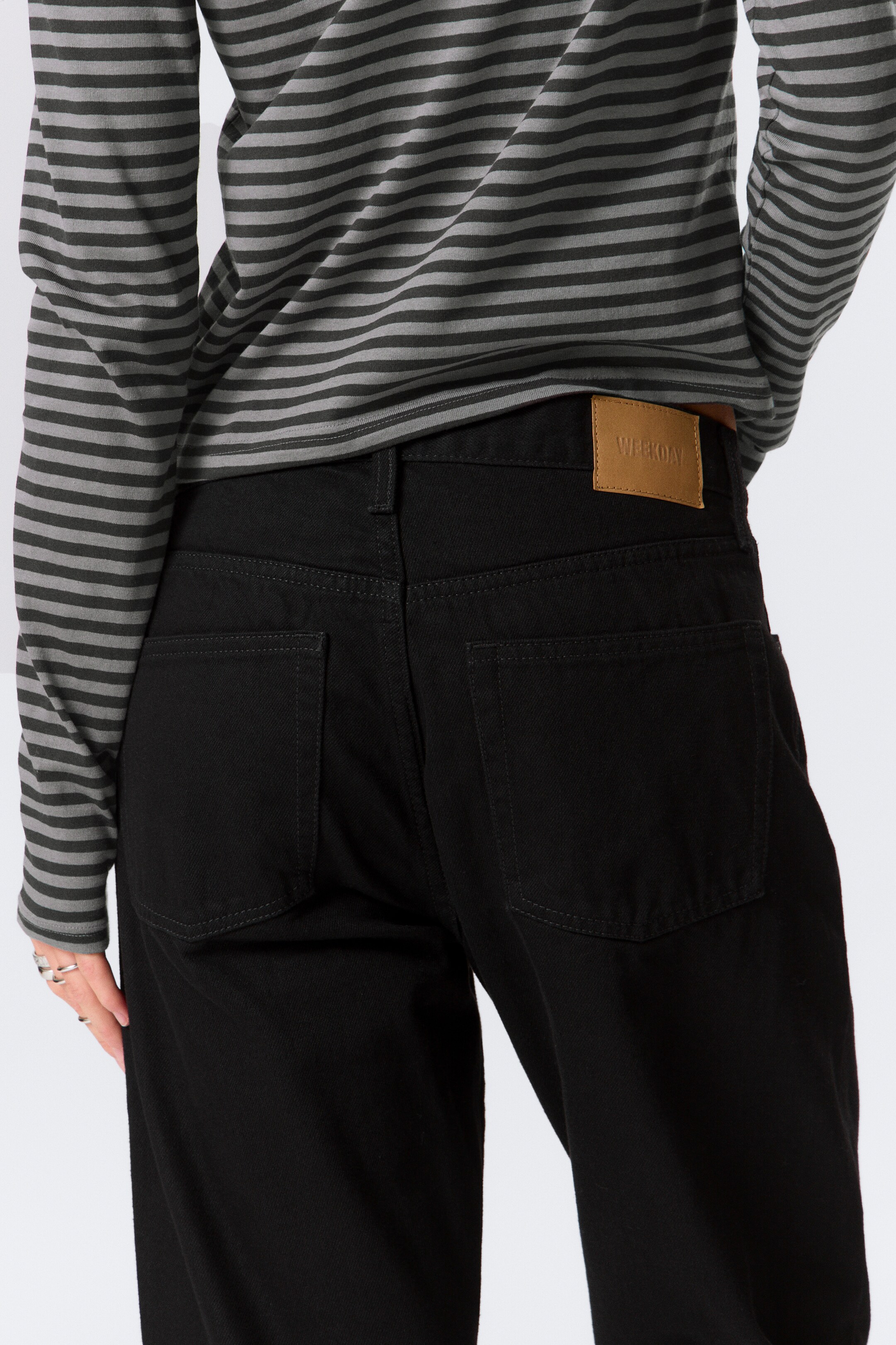 View larger image: Black jeans are being presented from the back, slightly angled to the right, revealing their relaxed fit. They are featuring two back patch pockets with visible contrast stitching and a tan leather-like brand patch embossed with "WEEKDAY" on the waistband, with a grey and dark grey horizontally striped long-sleeve top partially tucked in.