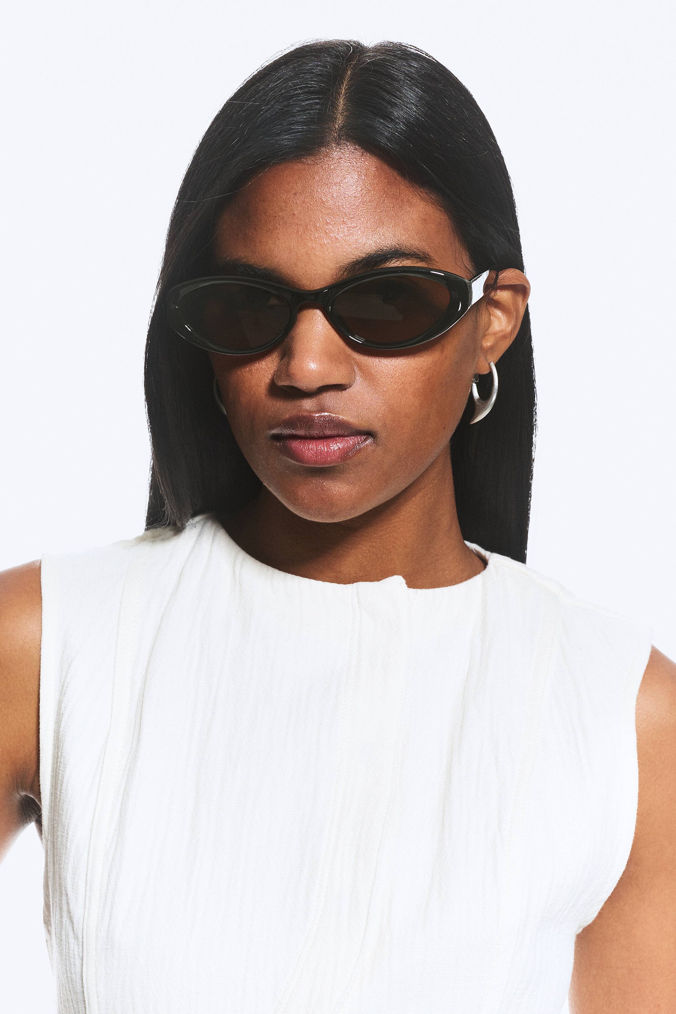 Größeres Bild ansehen: Ovale Sonnenbrille mit schwarzen Fassungen, dunklen ovalen Gläsern und silbernen Akzenten an den Bügeln wird vom Modell getragen.