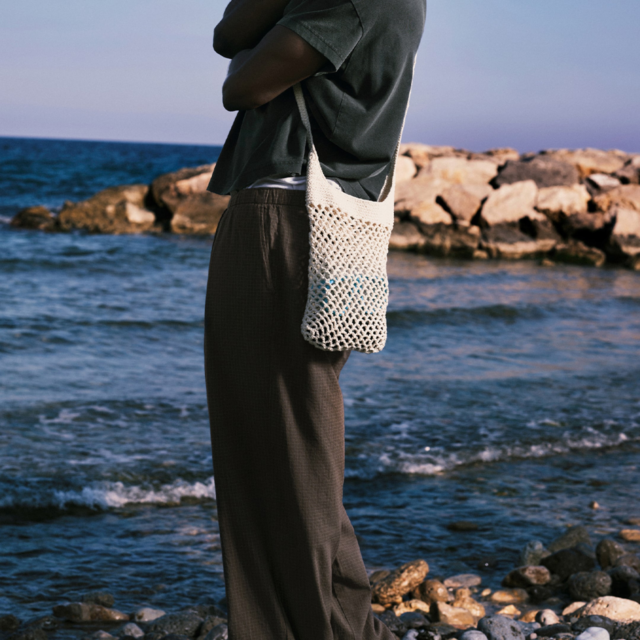 Model standing on a rocky beach with a netted bag, wearing a dark shirt and pants.