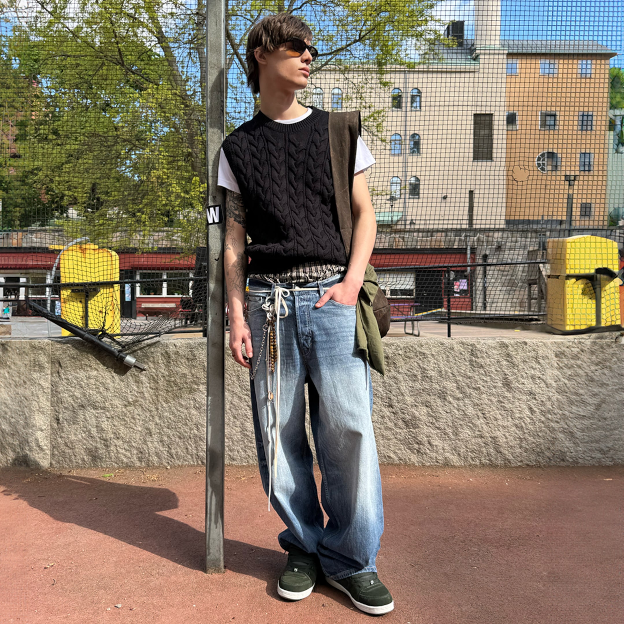 A person wearing sunglasses, a black sleeveless sweater, white shirt, and baggy jeans stands outdoors on a sunny day.