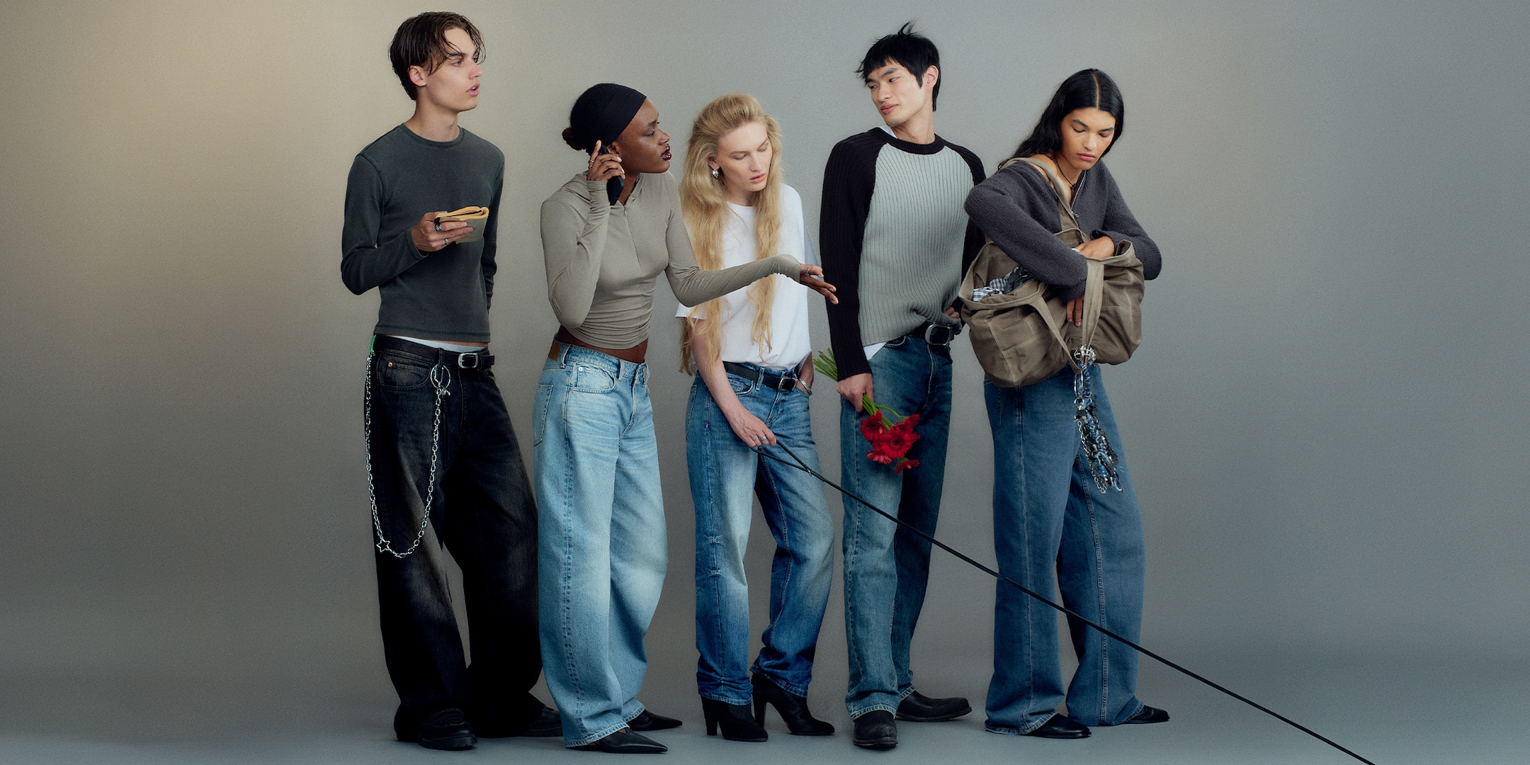 Five people in casual outfits stand against a neutral background, wearing jeans and tops, with one holding a red flower.