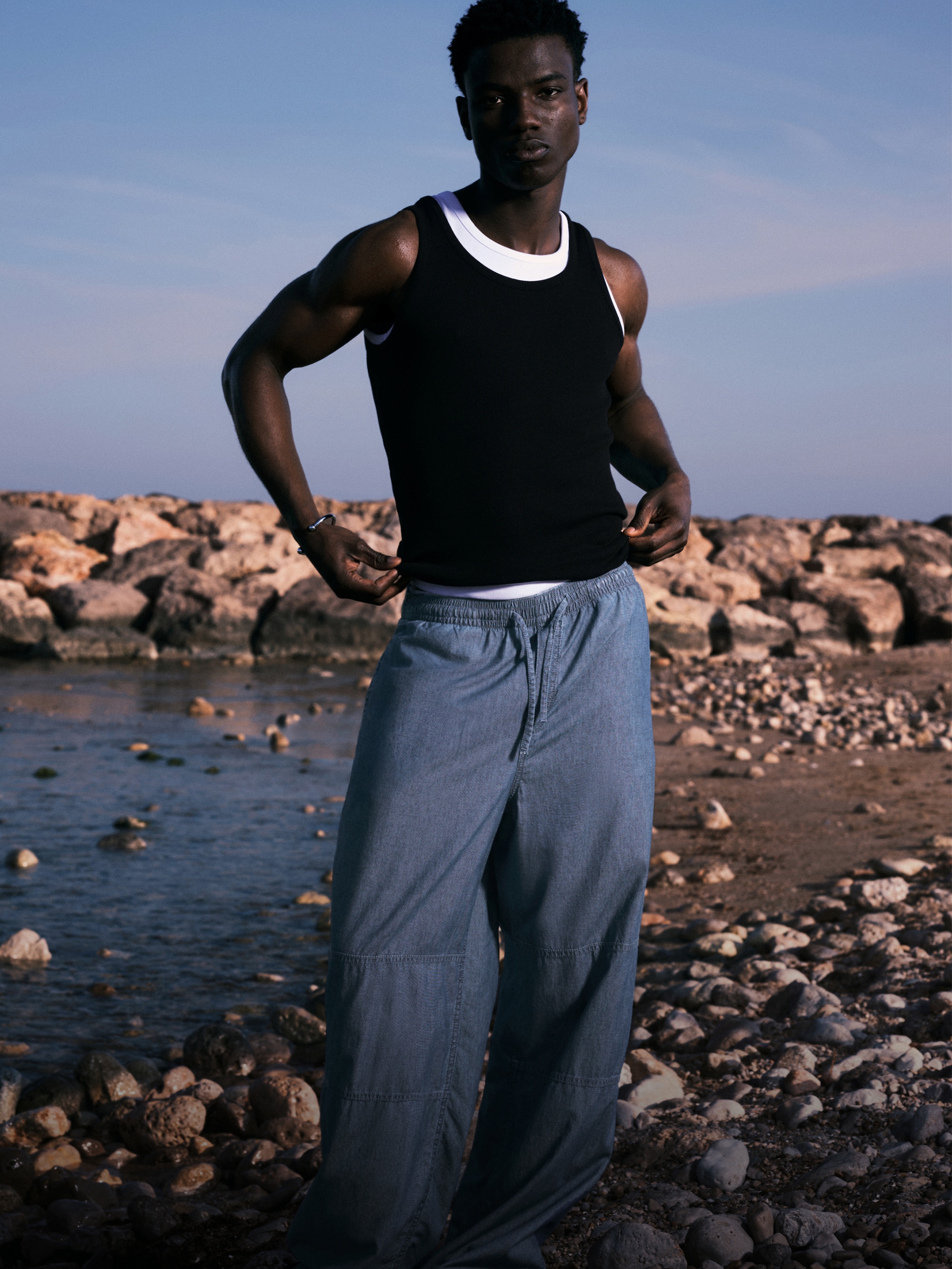 Model standing on a beach wearing a pair of blue trousers and a black tanktop